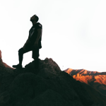 A person standing on a rocky mountain peak at sunset, silhouetted against a bright sky, with rugged cliffs on either side and sunlit mountains in the background.