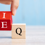 Hand placing red block with letters E and I on wooden Q block, symbolizing Emotional Intelligence over IQ in leadership.