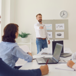Leadership coaching session in progress, with a coach presenting to a group of professionals seated around a conference table, reviewing charts and performance data.