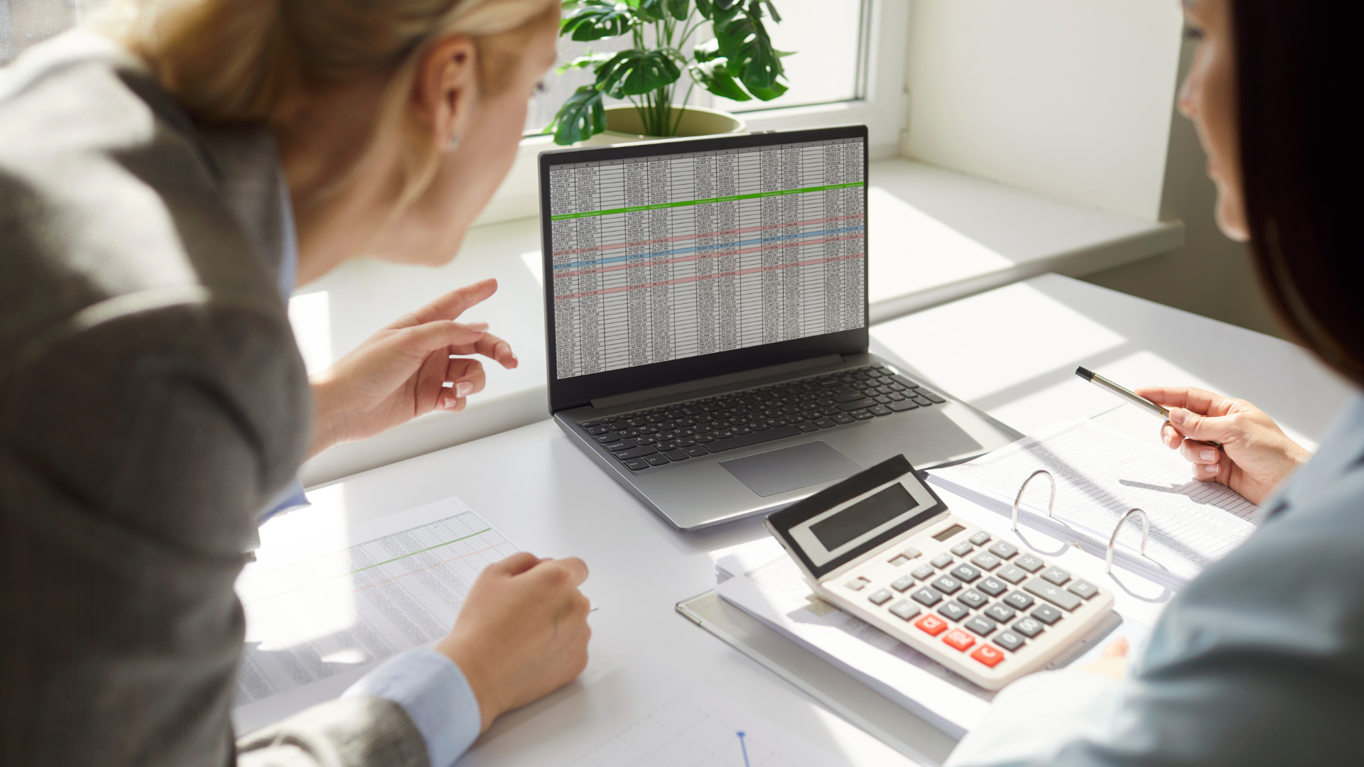Two business professionals analyzing management reports on a laptop with Excel spreadsheets, discussing data analysis and financial performance during a reporting meeting.