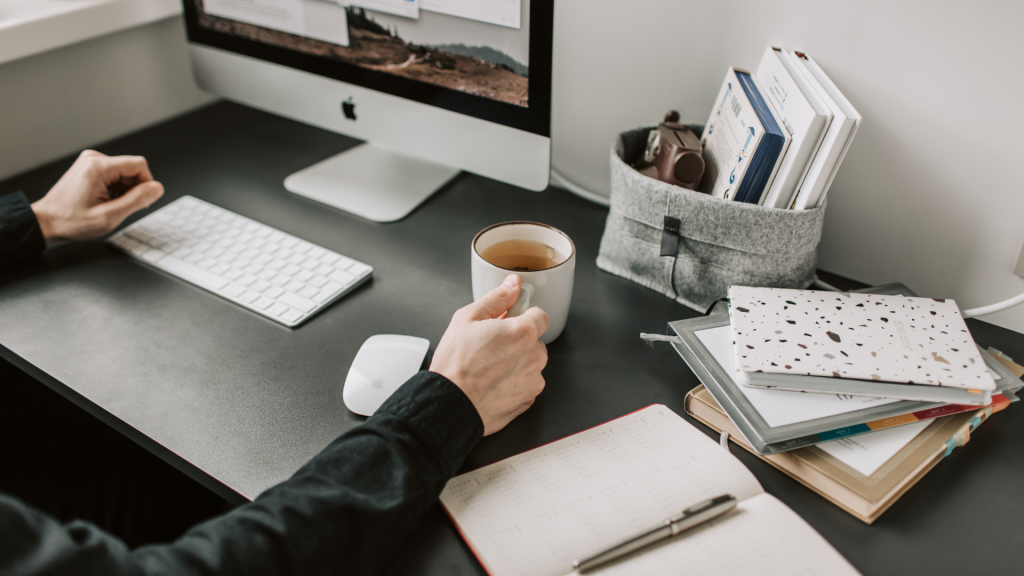 Person working at a minimalist office desk with computer, holding coffee, reviewing notes in a notebook, focused workspace setup for productivity, time management and professional work routine.
