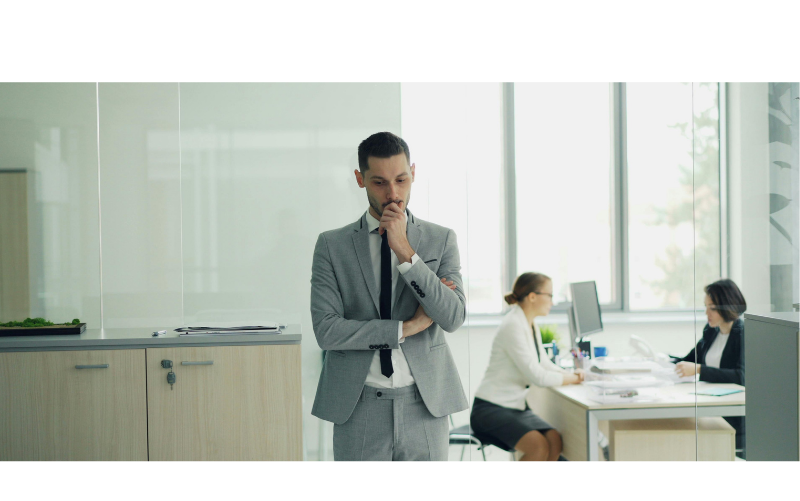 Thoughtful business leader reflecting on team communication challenges in a modern office environment, with colleagues working in the background, representing workplace communication, leadership, and team dynamics