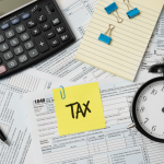 Flat lay of tax documents with calculator, alarm clock, pen, and sticky note labeled “TAX,” representing deadlines, financial planning, and tax preparation concept.
